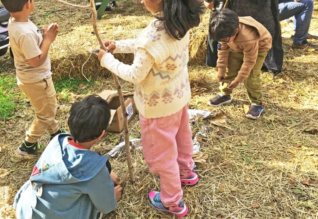 A photo of 4 small children building a den: tying sticks together, gathering straw, etc